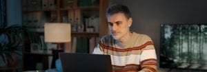 Muž v pruhovaném triku sedí u stolu a dívá se na notebook před sebou.